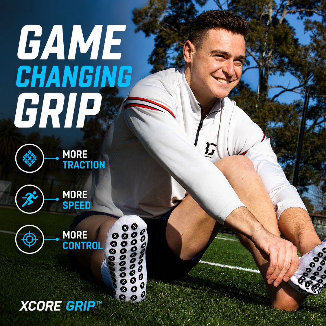 A man sitting on a grassy field outdoors wearing white athletic shoes with black grip dots and a white long-sleeve top. Large text reads 'GAME CHANGING GRIP' with feature callouts 'MORE TRACTION', 'MORE SPEED', and 'MORE CONTROL', plus 'XCORE GRIP'.