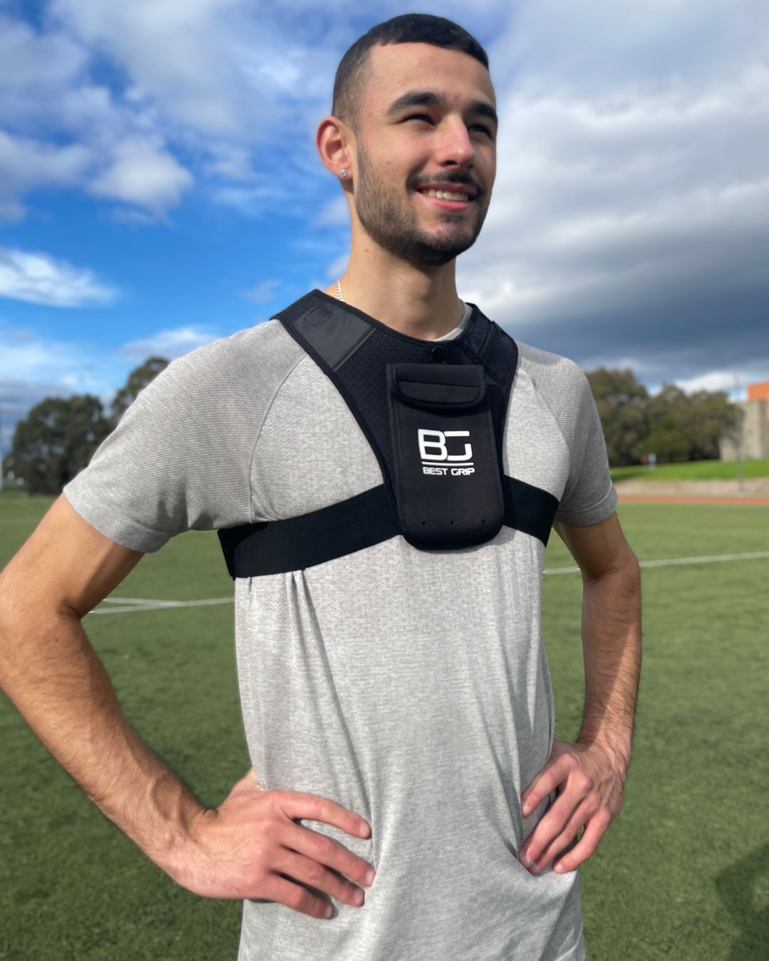 A man wearing a black training vest with a front pouch featuring the 'Besr Grip' logo, standing on an outdoor grass field under a cloudy blue sky.