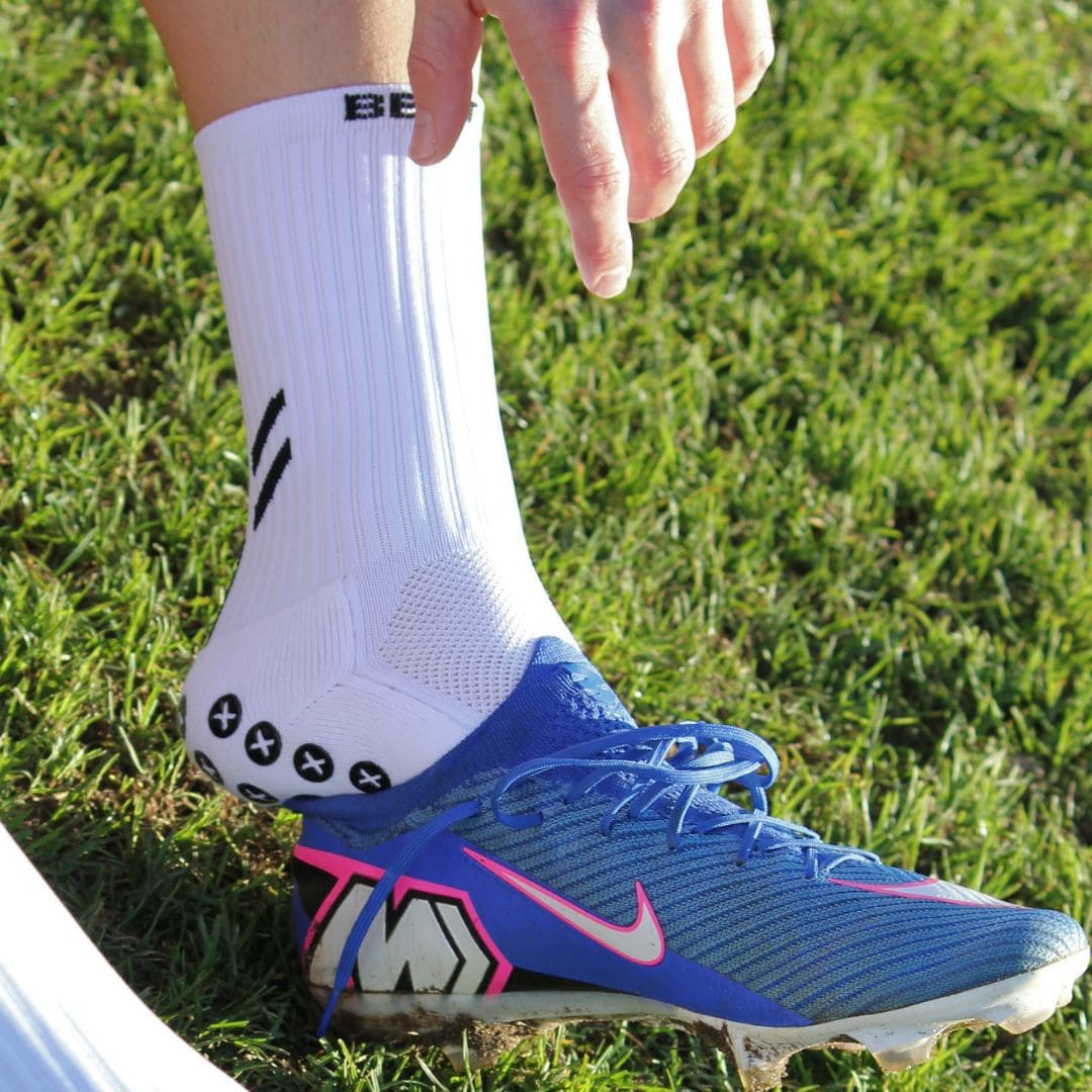 A close-up of a person's foot wearing a blue Nike soccer cleat and a white athletic sock on green grass. The sock has black logo details, and the shoe features a blue and pink Nike design.