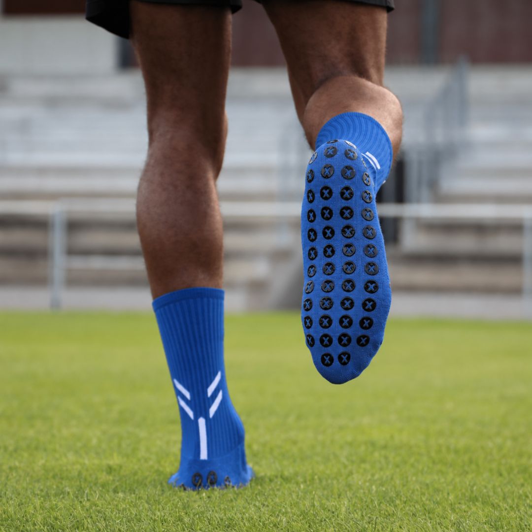 Athlete Wearing Blue Best Grip Crew Socks On A Grass Sports Field, With One Foot Raised To Reveal Black XCORE Grip™ Pads With Blue X Centres Across The Sole, Ribbed Cuffs And White Double Stripe Detailing, With Blurred Stadium Seating Behind.