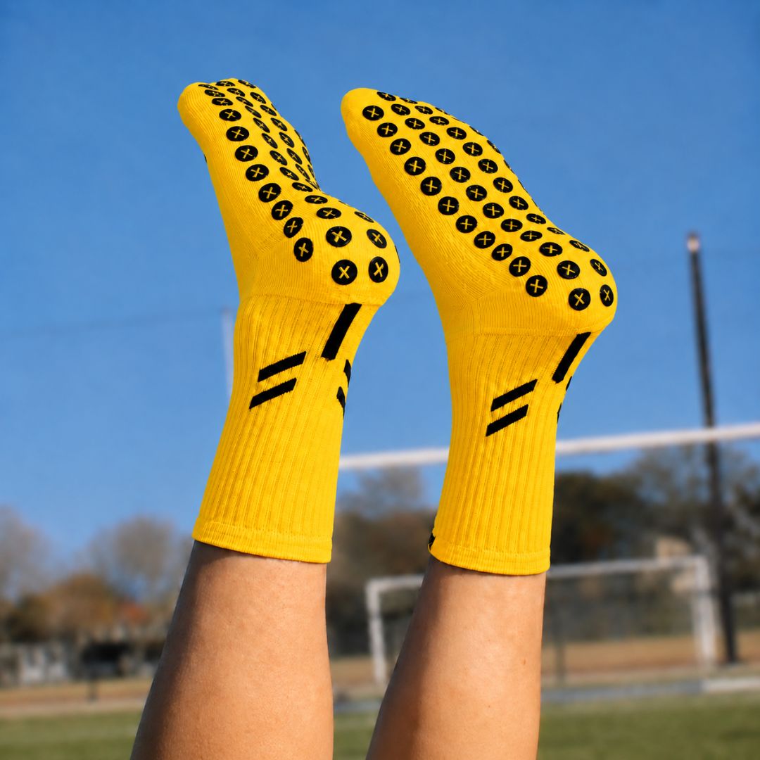 Pair of yellow Best Grip crew socks worn on raised legs outdoors, showing black XCORE Grip™ pads with yellow X centres across the soles, ribbed cuffs and black double stripe detailing against a blue sky and blurred sports field.