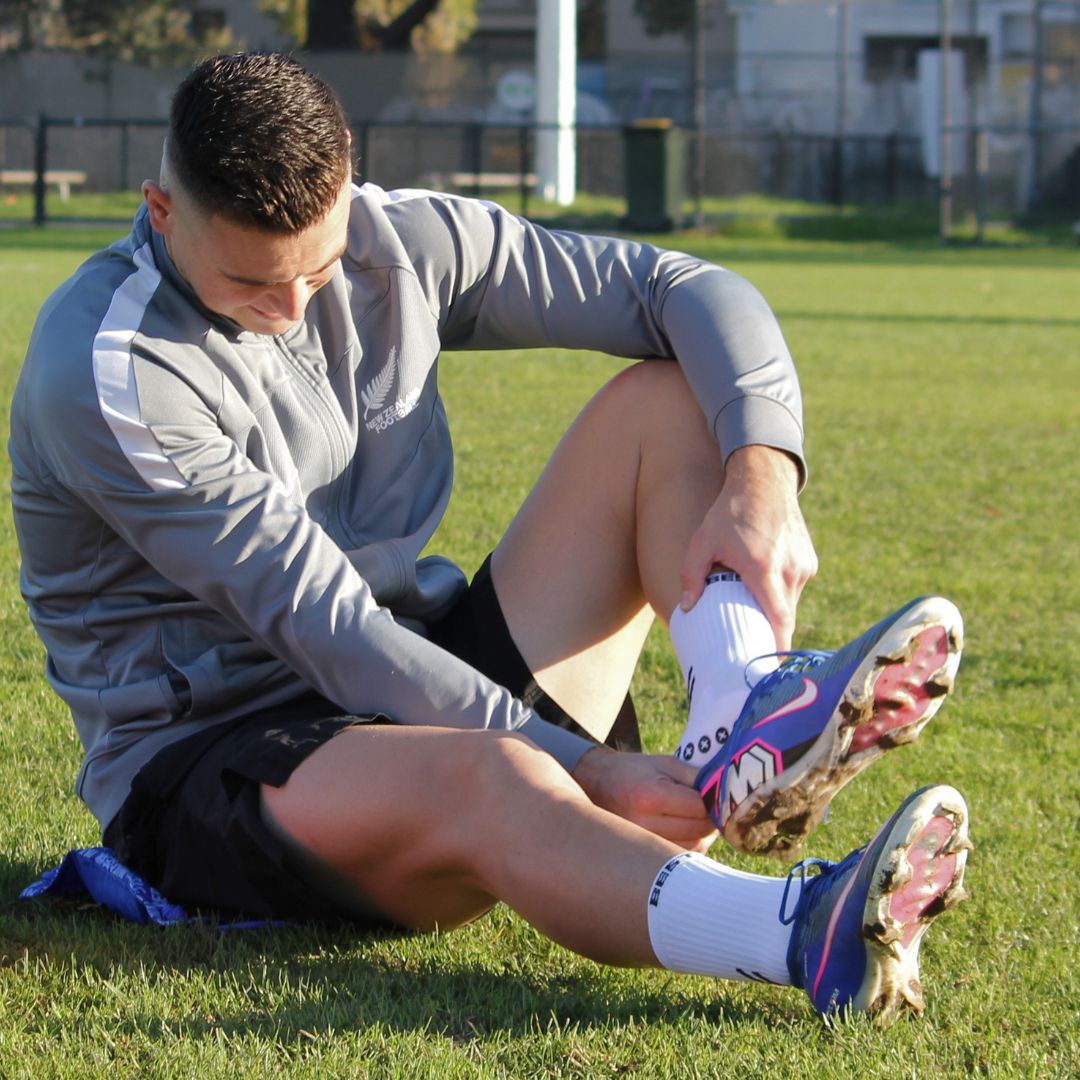 A man sitting on a grassy field outdoors, putting on a white athletic sock into his cleat. He is wearing a gray long-sleeve training top, black athletic shorts, and blue and pink cleats.